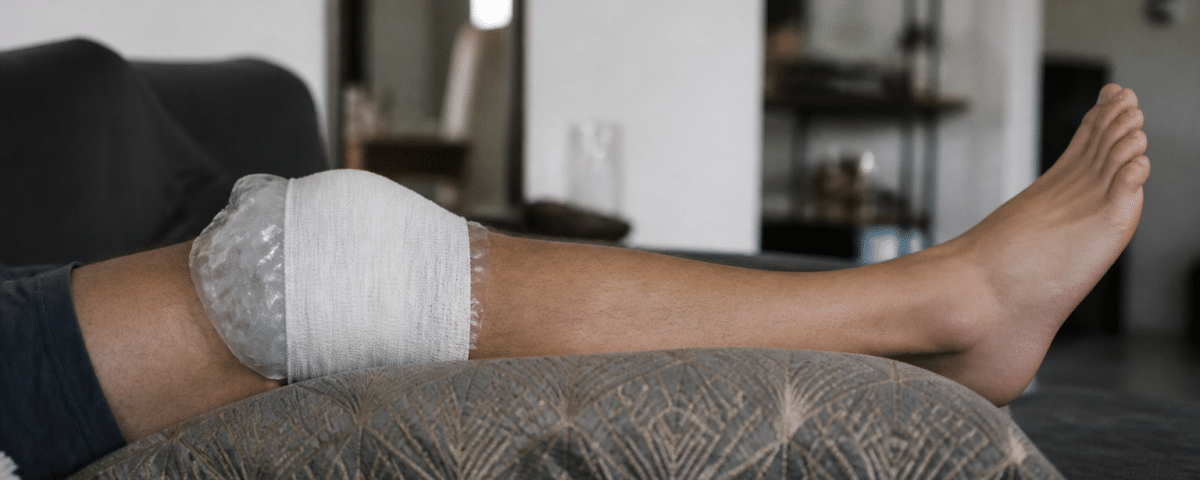 Genou bandé avec glaçon, jambe surélevée sur coussin.