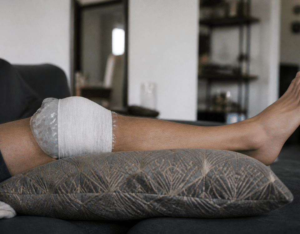 Genou bandé avec glaçon, jambe surélevée sur coussin.
