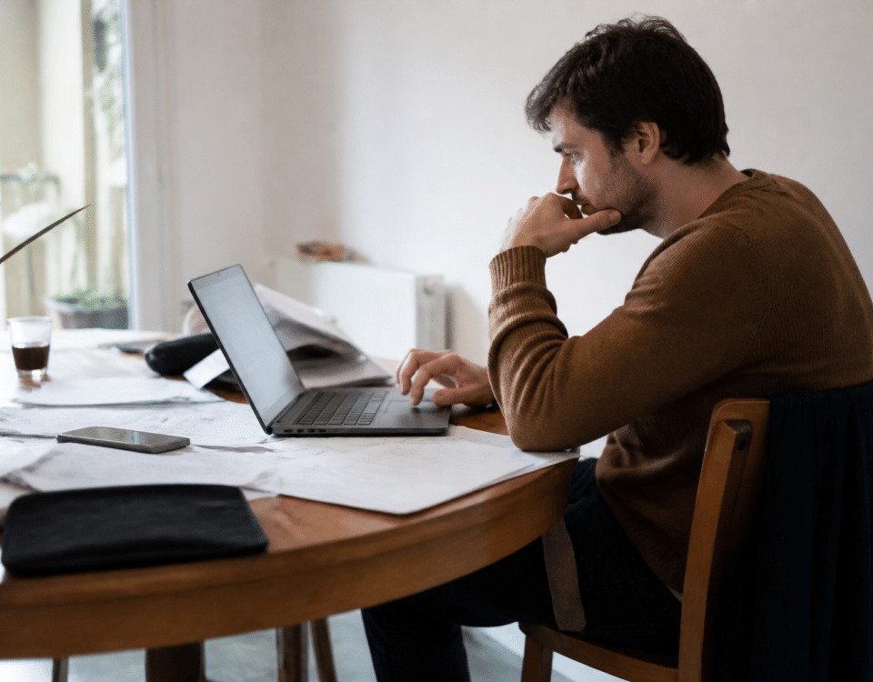 Homme assis consulte son ordinateur, documents eparpilles.